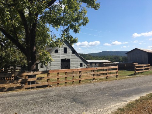 A rustic farm scene featuring a gray barn, wooden fence, and a clear blue sky with mountains in the background.