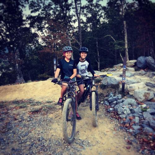 Two cyclists smiling on mountain bikes, surrounded by trees and rocky terrain on a trail.