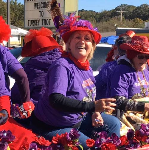 A smiling woman in a purple shirt and festive hat sits among others, celebrating at a lively outdoor event.