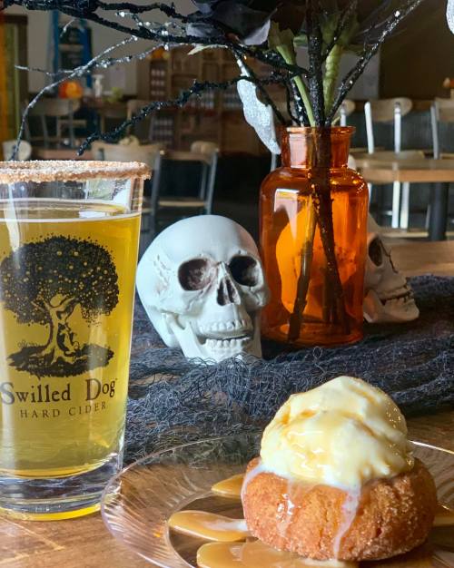 A glass of hard cider, a dessert with icing, and decorative skulls on a wooden table.