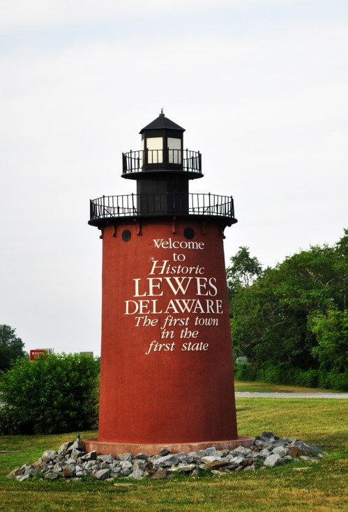 A red lighthouse with a black lantern, featuring text welcoming visitors to historic Lewes, Delaware, the first town in the first state.