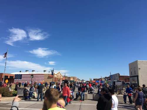 A bustling street fair with crowds of people, colorful tents, and a clear blue sky in a small town.