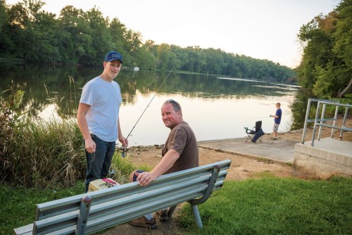 Two men sit on a bench by a river, one holding a fishing rod, while another fishes nearby. Trees line the water's edge.