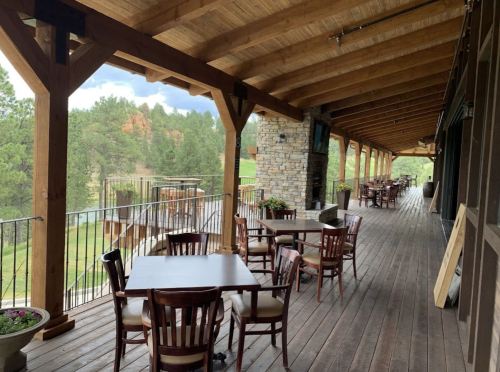 A wooden deck with tables and chairs overlooking a scenic landscape, featuring a rustic building and greenery.