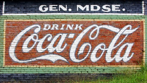 Vintage Coca-Cola advertisement painted on a brick wall, featuring bold white lettering on a red background.
