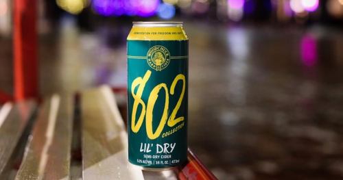 A can of 802 Lil' Dry cider on a bench, with a blurred colorful background.