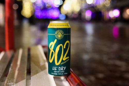 A can of 802 Lil' Dry semi-dry cider on a bench, with blurred festive lights in the background.
