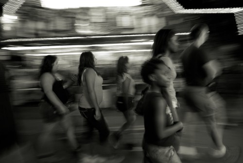 A blurred black-and-white image of people walking in a lively outdoor setting, with lights in the background.