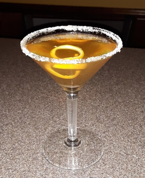 A cocktail in a clear glass with a salted rim and lemon twist garnish, resting on a textured surface.
