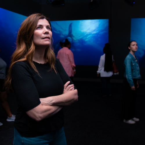 A woman stands thoughtfully in an art exhibit, surrounded by people and large ocean-themed displays.