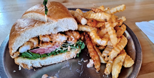 A crispy shrimp sandwich with lettuce and tomato, served with a side of crinkle-cut fries on a metal plate.
