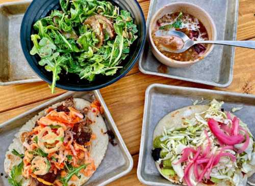 A spread of tacos with toppings, a bowl of arugula salad, and a small dish of soup on a wooden table.