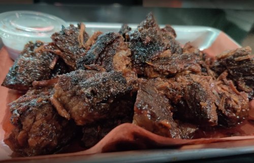 A close-up of a platter filled with dark, smoky pieces of beef, served with a small container of sauce.