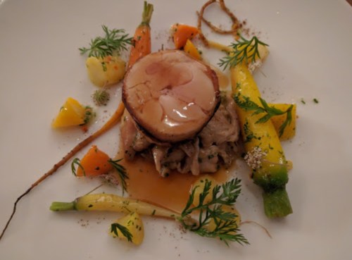 A gourmet dish featuring a round meat portion atop shredded meat, surrounded by colorful vegetables and garnishes.