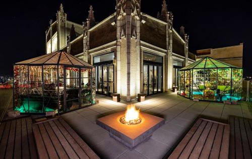 Rooftop terrace at night featuring two glass structures and a central fire pit surrounded by wooden benches.