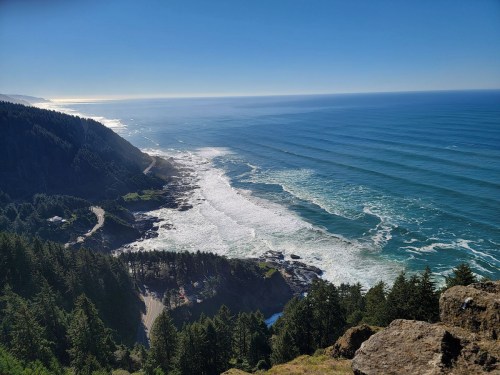 A scenic coastal view with waves crashing on the shore, surrounded by lush green forests and a clear blue sky.