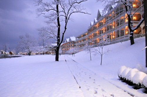 A snowy landscape with a large, lit building and trees, creating a serene winter scene at dusk.