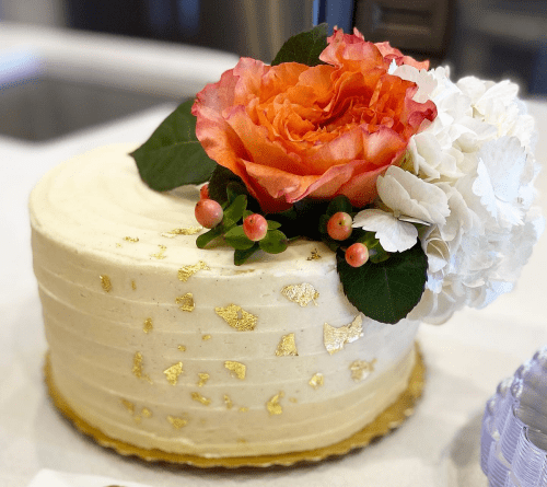 A beautifully decorated cake topped with a large orange rose, white flowers, and gold leaf accents.