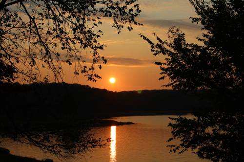 Sunset over a calm lake, with silhouettes of trees framing the scene and a warm orange glow reflecting on the water.