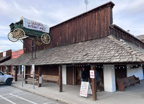 A rustic wooden building with a sign for "Hanna's Steak House" and a wagon above the entrance, featuring outdoor seating.