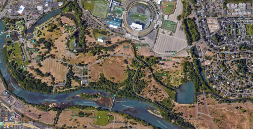 Aerial view of a park with a river, sports facilities, and surrounding neighborhoods.