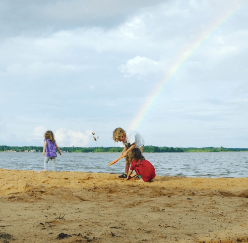 Children playing on a sandy beach with a rainbow in the background and a calm lake.