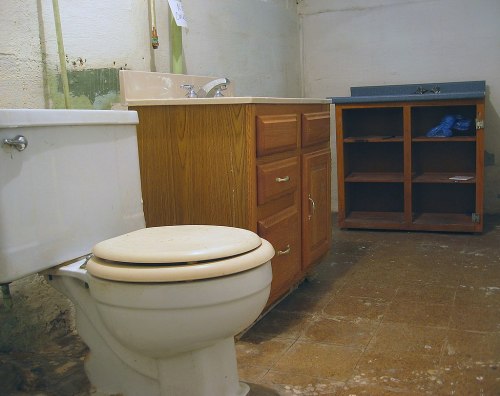 A small, unfinished bathroom featuring a toilet, wooden vanity with sink, and a cabinet against the wall.