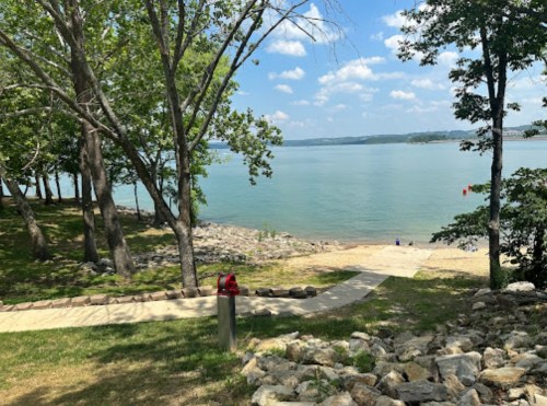 A serene lakeside view with trees, a sandy beach, and a clear blue sky, featuring a pathway leading to the water.