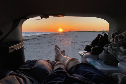A person relaxes in a car, watching the sunset over a beach, with feet visible and belongings in the background.
