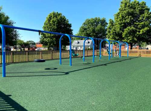 A sunny playground with blue swings and a tire swing, surrounded by green grass and trees.