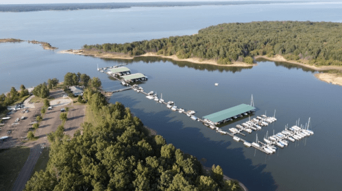 Aerial view of a marina with boats docked, surrounded by trees and calm water under a clear sky.