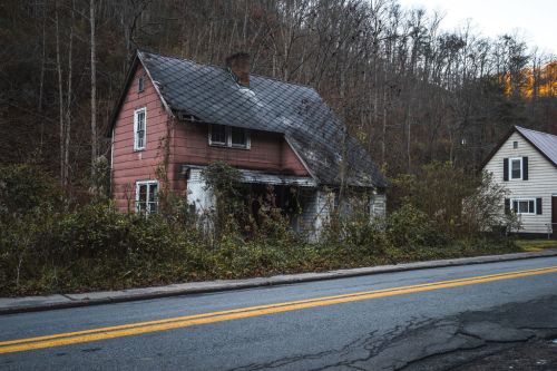 Abandoned house with a weathered pink exterior, surrounded by overgrown vegetation, along a quiet road.