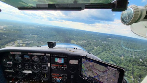 Cockpit view from a small aircraft, showing flight instruments and a GPS map over a green landscape below.