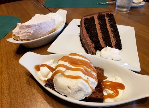 Three desserts on a wooden table: a slice of chocolate cake, a brownie with ice cream and caramel, and a slice of pink pie.