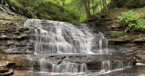 A serene waterfall cascading over rocky steps, surrounded by lush green trees and a peaceful forest setting.