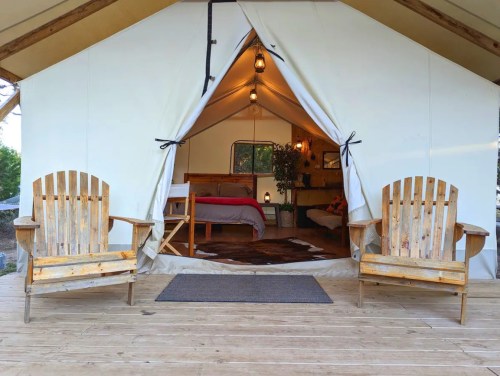 A cozy glamping tent entrance with two wooden chairs, a bed inside, and warm lighting.