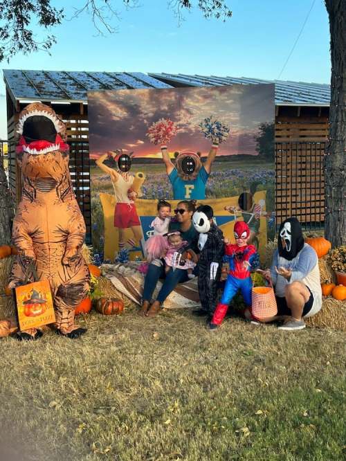A family poses in costumes, including a dinosaur and Spider-Man, in front of a festive fall backdrop with pumpkins.