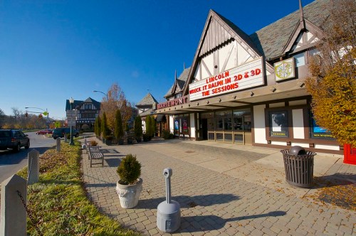 A quaint theater building with a marquee, surrounded by trees and a clear blue sky. Sidewalk and street visible.
