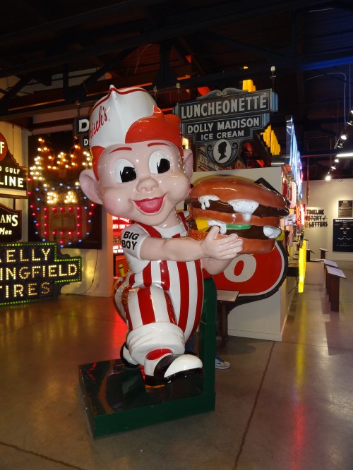 A large, cheerful statue of a boy in a striped outfit holding a sandwich, surrounded by vintage neon signs.
