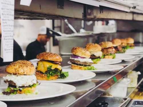 A row of gourmet burgers on plates, each with different toppings, lined up in a busy kitchen.