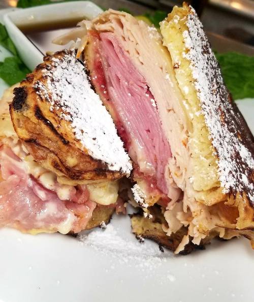 A close-up of a sandwich filled with layers of ham and turkey, dusted with powdered sugar, served with dipping sauce.