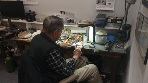 An elderly man works at a cluttered desk, focused on a small project with tools and materials around him.