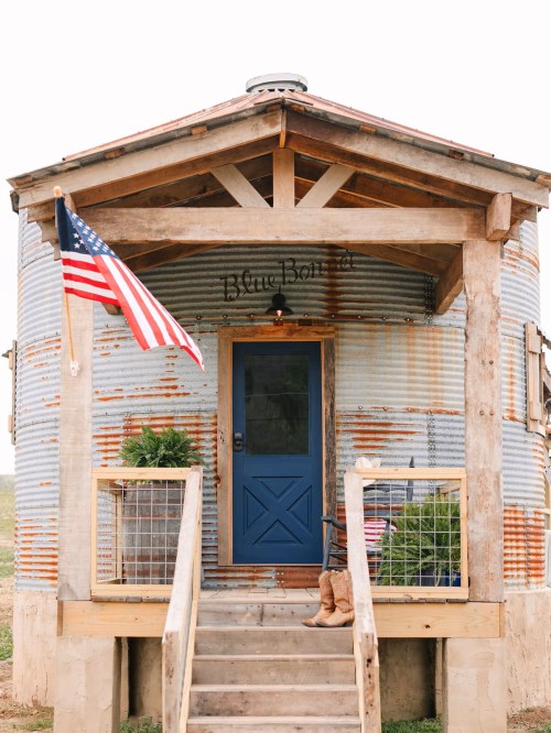 A rustic metal building with a blue door, wooden steps, and an American flag, surrounded by greenery.