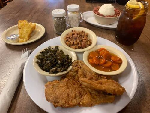 A plate of fried chicken with collard greens, black-eyed peas, carrots, and cornbread, alongside a drink and dessert.