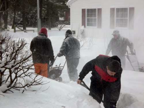 A group of people shoveling snow in a snowy yard, with a house visible in the background. Snow is blowing around them.