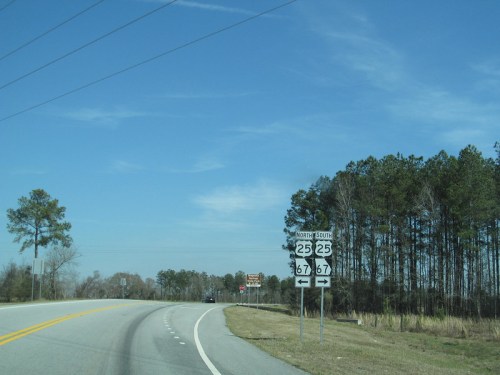 Road sign indicating routes 25 and 67, with a clear blue sky and trees lining the roadside.