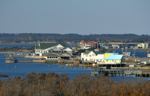 Coastal view of a small town with colorful buildings along the water, surrounded by trees and distant hills.