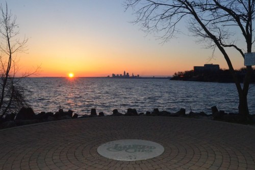 Sunset over a lake with a city skyline in the distance, framed by trees and a circular stone path in the foreground.