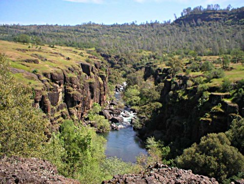 A serene river flows through a lush canyon, surrounded by green hills and rocky cliffs under a clear blue sky.