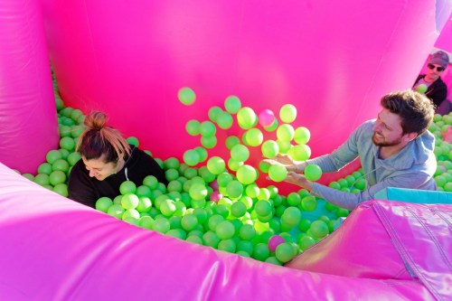 Two people play in a colorful ball pit filled with green balls, surrounded by bright pink inflatable walls.